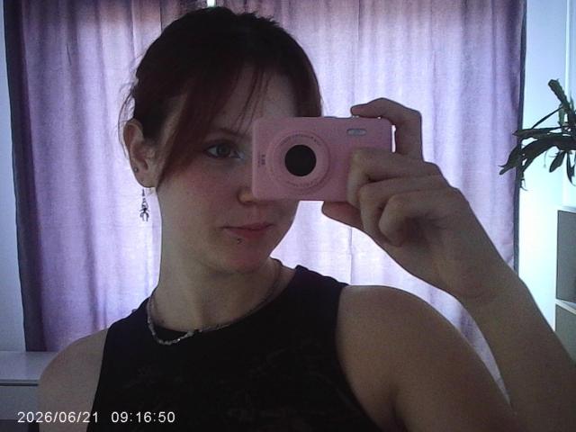 PatSanabria, con cabello rojo oscuro, sosteniendo una cámara rosa, vistiendo una blusa negra con encaje, mostrando un piercing en la nariz y un collar de plata, en un selfie frente a un espejo..