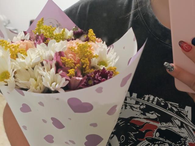 Genesiss tiene in mano un bouquet di fiori bianchi, rosa e viola con elementi verdi gialli, indossa una maglietta nera con una grafica e tiene in mano un telefono rosa con unghie rosse e nere..