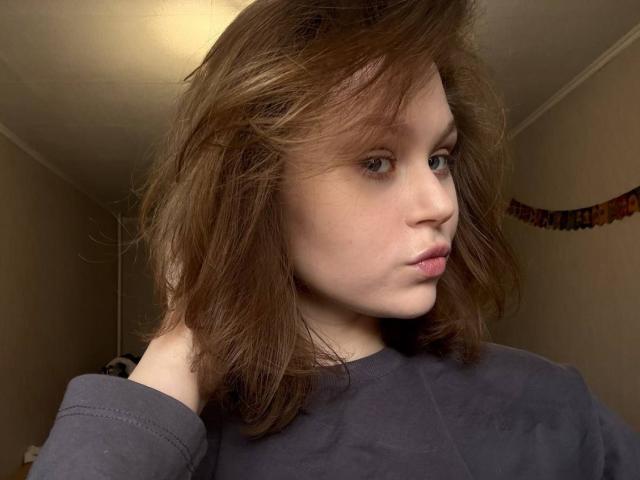 YungQuiller ha la pelle chiara, i capelli castano ondulati fino alle spalle, e indossa una camicia grigia con una spalla scoperta, guardando di lato con un leggero broncio..