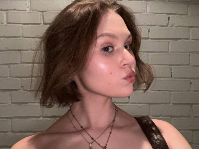 YungQuiller, con cabello castaño corto, labios fruncidos, maquillaje sutil, piel brillante, vistiendo una correa marrón y dos collares, frente a una pared de ladrillos blancos..