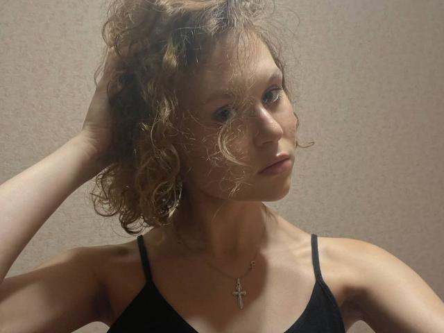 YungQuiller mit lockigem, braunem Haar, Hand im Haar, trägt ein schwarzes Tanktop, eine kleine Kreuzkette, blickt zur Seite, leichtes Schmollen, blasse Haut, dezentes Make-up, weiches Licht..