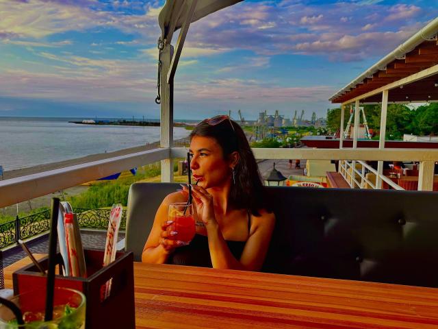 LakiaRentz, with tits visible, sips a red drink, sitting in a black booth, wearing a strapless top, sunglasses on head, wooden table, ocean view, cloudy sky, and city.