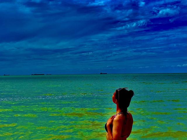 LakiaRentz with brown skin and black hair in a bikini, standing in greenish-blue water, looking at a dark blue, cloudy sky, with a few boats on the horizon.