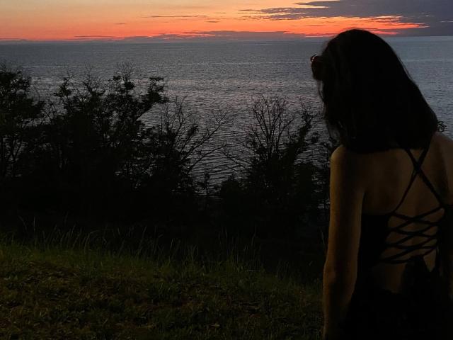 LakiaRentz with dark hair, back to camera, wearing a black dress with crisscross straps, standing on grassy hill, watching sunset over dark ocean, silhouetted against orange sky.
