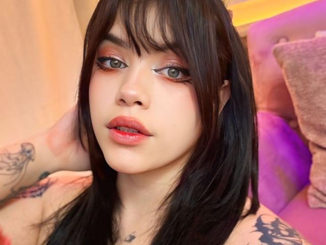 AryButty with pale skin, black hair, bangs, and red lipstick, looking at the camera, showing tattoos on her arm and shoulder, with pink and purple pillows in background.