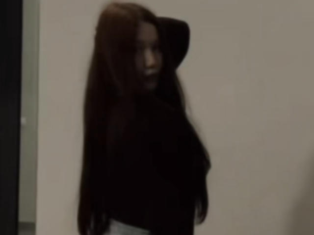 Zita Sutterfield, com cabelos longos e castanho-escuros, vestindo uma blusa preta, tem um braço levantado, exibindo suas nádegas e um pequeno volume visível no seio. Imagem borrada e granulada..