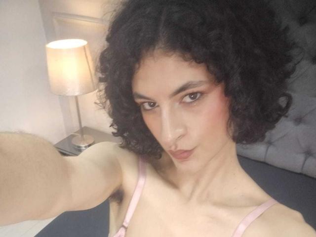 RoxyGraff hat lockiges, schwarzes Haar, hellbraune Haut und ein leichtes Lächeln. Sie trägt ein pinkes Top mit dünnen Trägern und hat sichtbare Achselhaare. Sie macht ein Selfie in einem sanft beleuchteten Raum..