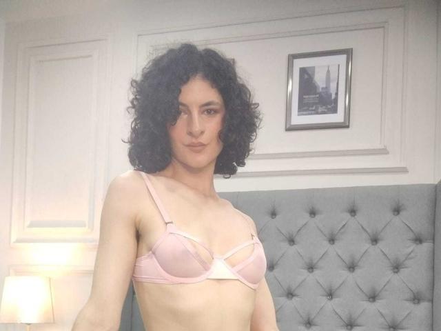 RoxyGraff, com cabelo preto encaracolado, usando um sutiã rosa, mostrando seios pequenos, em pé em frente a uma cabeceira cinza com textura, com um abajur suave ao fundo..