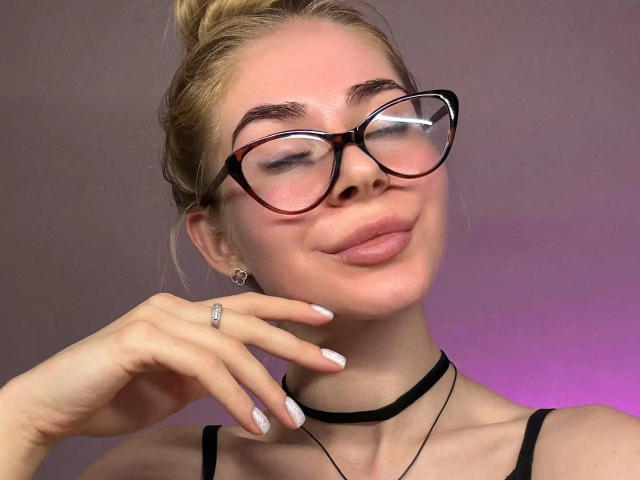 BarbaraBlum with glasses, blonde hair in a bun, black choker, and pink lips, smirking, hand near face, ring on finger, white nails, purple light in background.