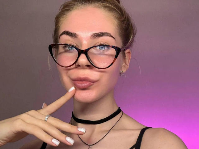 BarbaraBlum with blue eyes, black glasses, and a slight smile, touching her chin with her finger, wearing a black choker and a black top. Her nails are white.