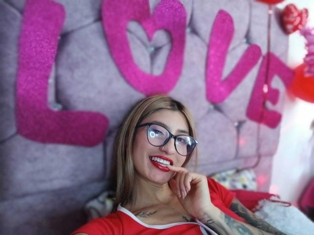 Kaiix, con gafas, lápiz labial rojo y tatuajes, mordiéndose el dedo, sonriendo, vistiendo una camisa roja, sentada sobre un cabecero gris con capitonado..