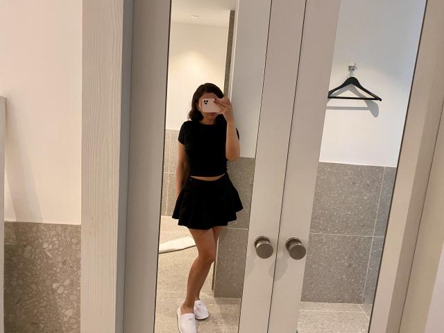 Dionisia's mirror selfie showing her black crop top, flirty skirt, white slippers, and holding a phone with a pink case, covering her face. Her ass is slightly visible.