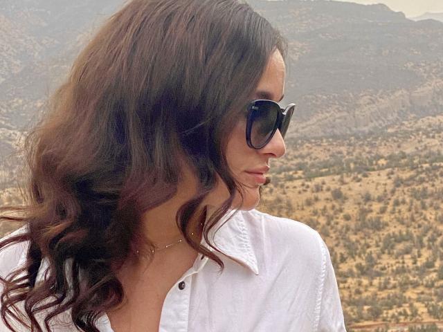 Dionisia with wavy brown hair, wearing black sunglasses and a white button-up shirt, standing with a calm expression. Boobs partially visible under shirt. Outdoor setting.
