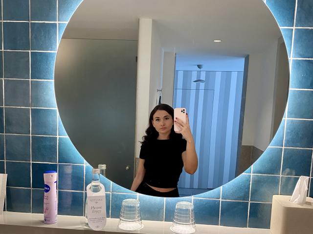 Dionisia with dark hair, black top, and black pants, taking a mirror selfie in a bathroom with blue tiled mirror. Her phone is pink, and she's holding it in her right hand.