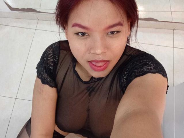 MelanieDiiaz, with red hair and purple eyeliner, stares up at the camera, tongue slightly out, wearing a sheer black lace top, revealing her small tits and nipples.