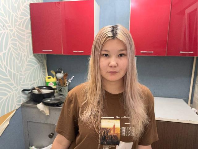 Selena Zappulla ha i capelli biondi e lisci, la pelle chiara e indossa una maglietta marrone. Si trova in una cucina con mobili rossi. Guarda direttamente la telecamera con un'espressione neutra..
