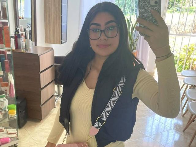 NinaRouse with long black hair, glasses, and a pink purse, taking a selfie in a salon, wearing a beige top and black vest, smiling at the camera.