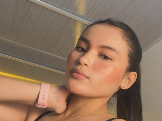 MiiaPalmer's face with pink skin tone, brown eyes, and dark hair in a ponytail, wearing a pink smartwatch, earrings, and a gold necklace, looking slightly to the side.