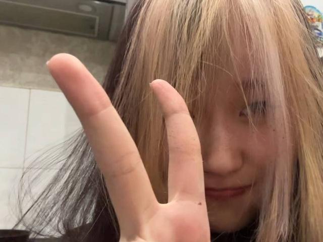 JungMcgranor with blonde and black hair, making a peace sign with her right hand, smiling slightly, in a kitchen. Her face is partially hidden by her long hair.