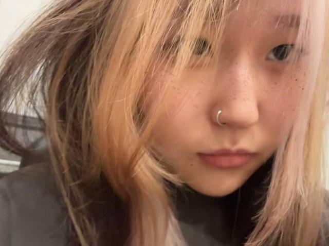 JungMcgranor has dirty blonde hair, a nose ring, and freckles on their face. Their lips are slightly parted in a neutral expression. The image is a selfie.