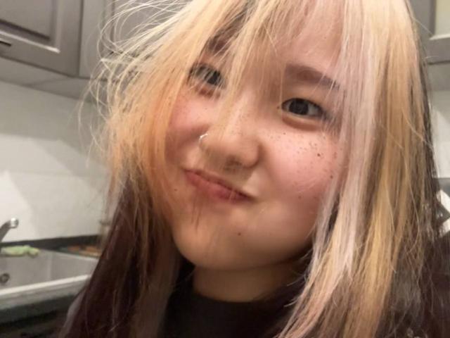 JungMcgranor with blonde and dark brown hair, nose piercing, freckles, and a slight smile in a kitchen. Her tits are not visible, just her face.