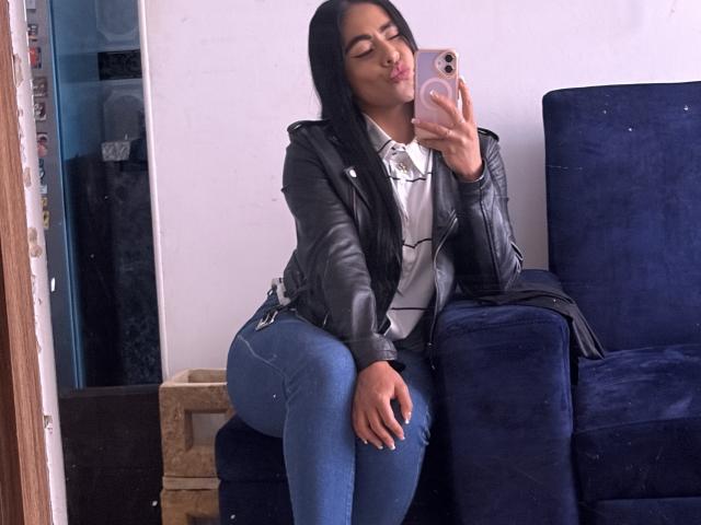 StellaBeckker, avec ses longs cheveux noirs, ses lèvres pulpeuses, sa veste en cuir noir, sa chemise blanche et son jean bleu, est assise sur un canapé bleu foncé, tenant un téléphone rose et embrassant son reflet. Ses fesses sont proéminentes..