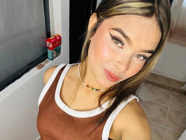 ChloeVixen with light brown tank top, white trim, small breasts, blonde highlights, green eyes, black eyeliner, pink lips, necklace, standing in front of window, tile floor.
