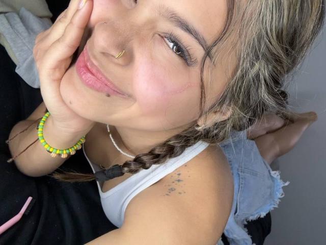 ChloeVixen with braided brown hair, nose ring, and lip piercing, smiling, touching her cheek, wearing a white tank top and yellow beaded bracelet, showing a small tattoo on her arm.