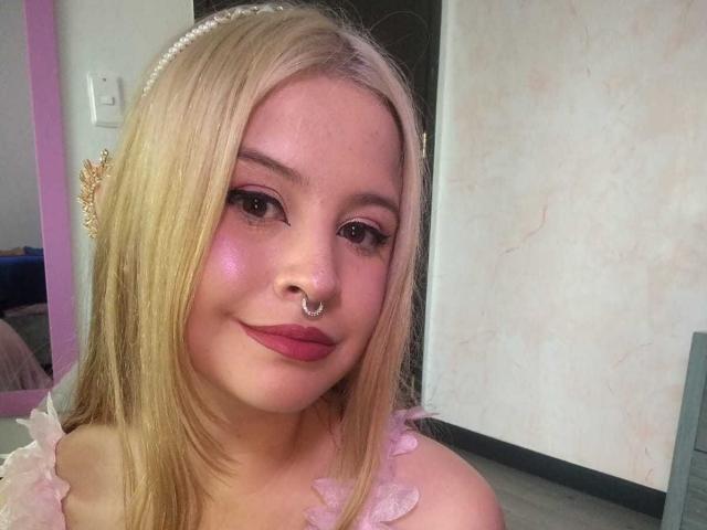 MelaniBecker with blonde hair, pink makeup, and nose ring, wearing a pearl headband and pink frilled top, smiling at the camera. Her tits are not visible.