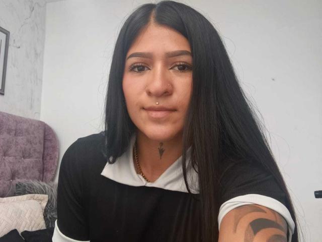 LuciadelaVega has long black hair, a nose piercing, and a tattoo on her right arm. She's wearing a black and white top, smiling slightly, with a necklace.
