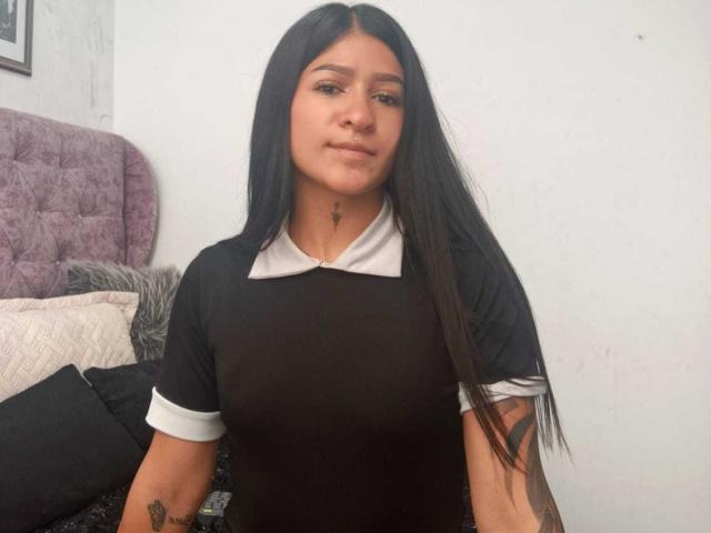 LuciadelaVega with long black hair, wearing a black shirt with white collar, small tits, tattoo on right arm, light brown skin, subtle smile, and small nose ring.
