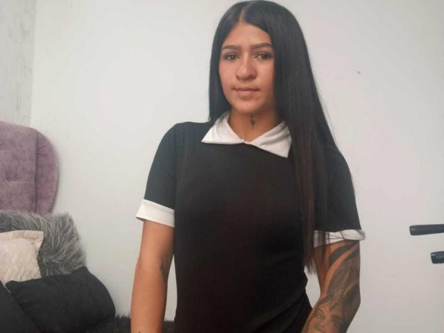 LuciadelaVega with long black hair, wearing a black and white nurse outfit, tattooed arm, and slight smile, standing in a room with a purple chair and gray pillows.