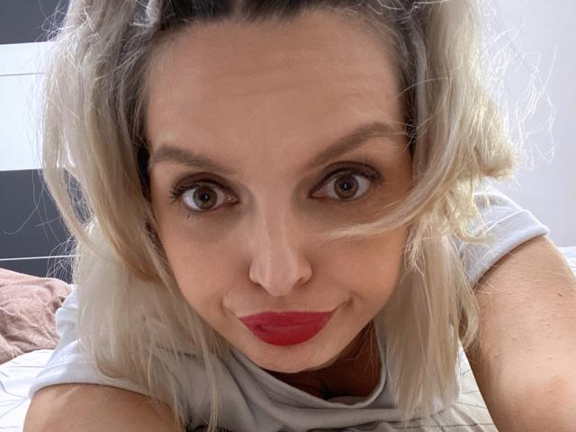 DouceAmelia with blonde hair, red lipstick, and brown eyes, staring directly into the camera, smiling slightly, wearing a white shirt, in a selfie-style shot.