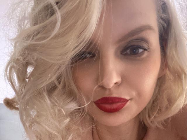O rosto de DouceAmelia, com cabelos loiros ondulados, batom vermelho e um olho ligeiramente fechado, exibindo seus lábios cheios e maquiagem discreta. Seus seios não estão visíveis..