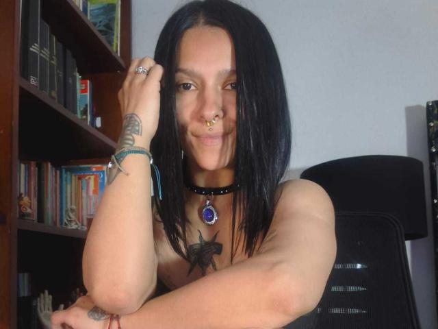 AmelieBlake with black hair, nose ring, and choker, smiling at the camera, topless, showing a tattoo and a blue gem necklace, arm resting on chair.