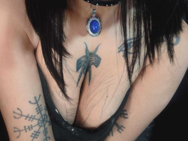 De borsten van Amelie Blake, versierd met een zwarte choker met een blauwe steen, laten tatoeages zien: een kruis op haar borst en ingewikkelde ontwerpen op haar armen..
