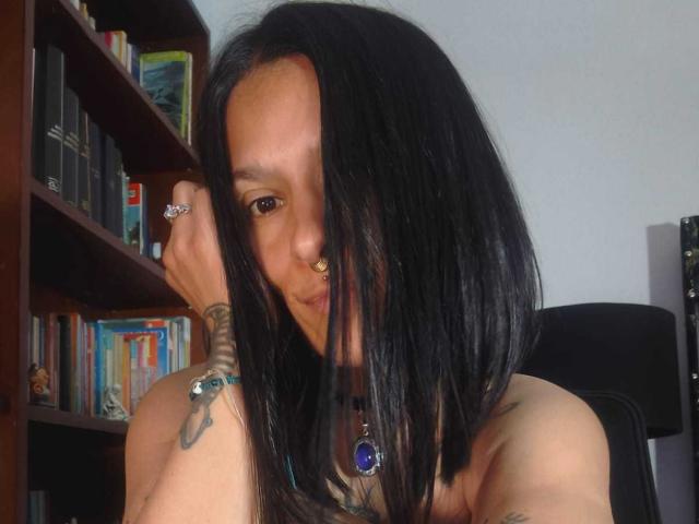 AmelieBlake with black hair, nose ring, and tattoos, wearing a purple gem necklace, holding her hair, showing her bare shoulders, and smiling slightly. Bookshelf in the background.