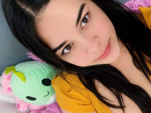 AuroraSantorini has dark hair, brown eyes, and a slight smile. She's wearing a yellow top with a green stuffed animal and pink pillow in the background.