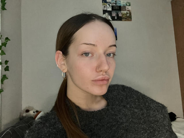 JamesAughenbaugh trägt einen Nasenring, hat kleine Brüste und einen grauen Pullover, einen Pferdeschwanz und hellrosa Lidschatten, und blickt mit leichtem Schmollen in die Kamera..