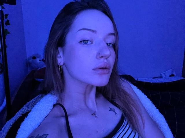 JamesAughenbaugh with blue-tinted skin, long brown hair, nose ring, gold earrings, star tattoos on chest, striped top, white and black robe, looking at camera with slightly parted lips.