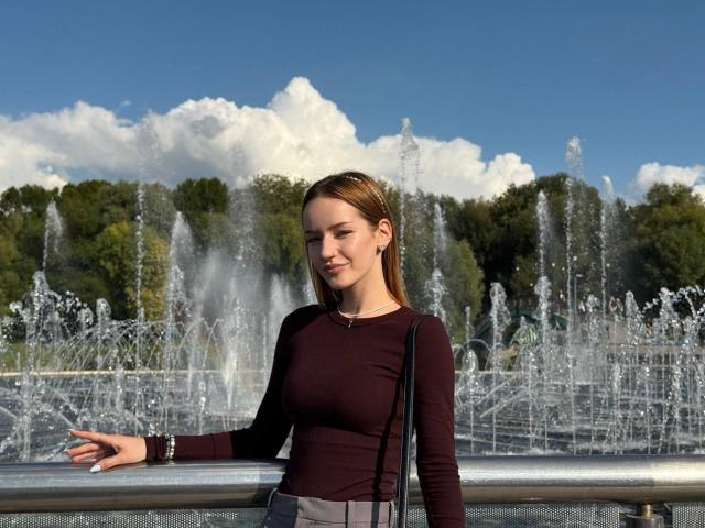 JamesAughenbaugh stands in front of a fountain, wearing a tight maroon top, gray skirt, and black shoulder bag, with a small smile and brown hair. Water jets rise.