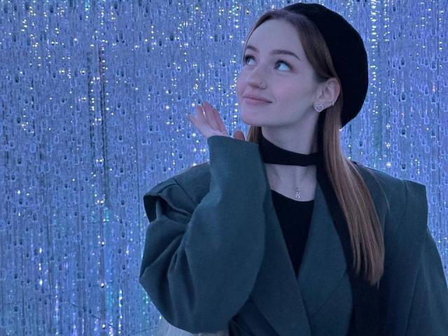 JamesAughenbaugh with long brown hair, wearing a black beret, green oversized jacket, black choker, and earrings, looking up with a slight smile, holding her hand near her face.