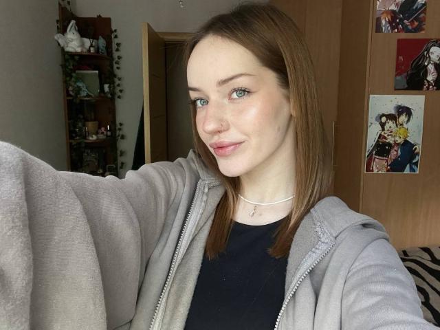 JamesAughenbaugh with pale skin, green eyes, brown hair, light pink lipstick, wearing a gray zip-up and black shirt, small nose, necklace, smiling, holding out arm.