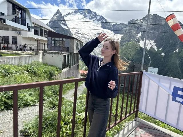 JamesAughenbaugh standing on a balcony, wearing a navy hoodie and green pants, hand in hair, looking up, with snowy mountains and a house in the background, sunny day.