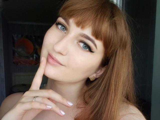 VioletAn has red hair, blue eyes, and a cute smile with a finger touching her chin. She's wearing gold earrings and has white nail polish.