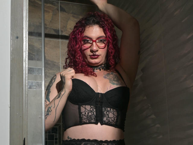 Skarleth69 met krullend rood haar, rode bril, een zwarte kanten bustier, tatoeages, een choker en een neusring, met haar arm omhoog, kijkend naar de camera, in een zwak verlichte douche..