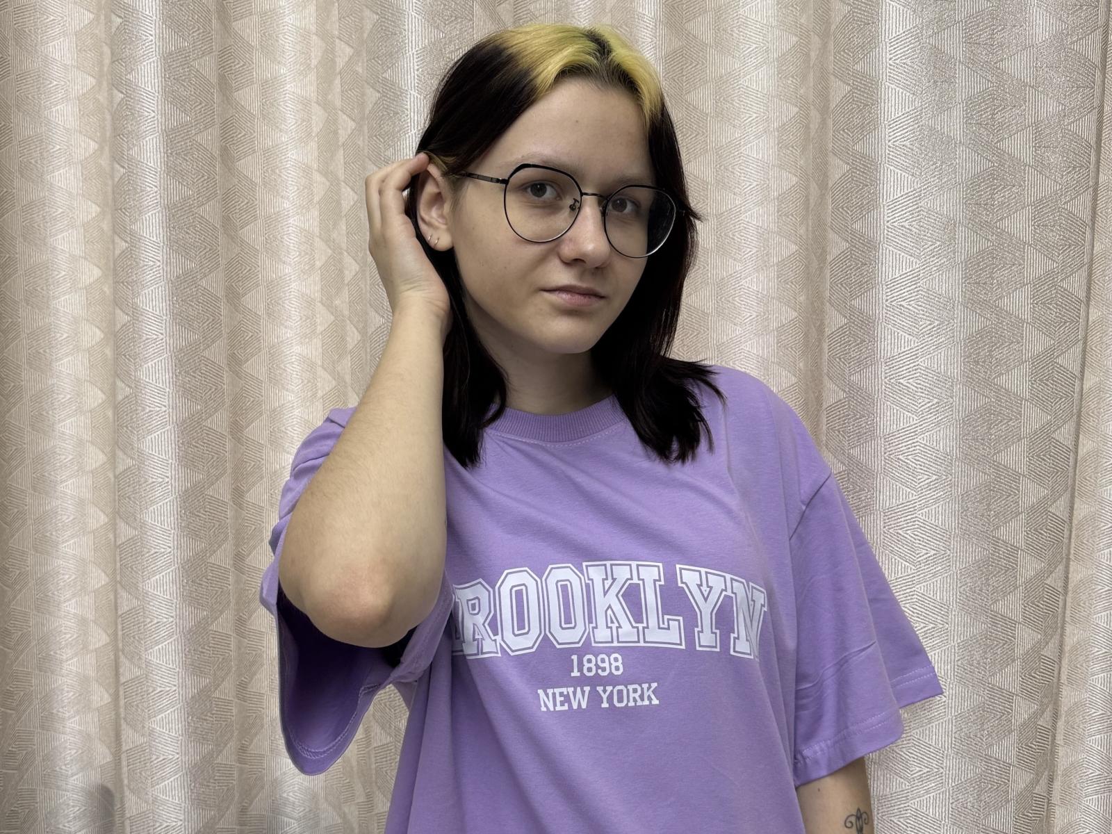 DortheaCalixtro with black and blonde hair, wearing glasses, and a purple "ROOKLYY 1999 NEW YORK" shirt, touching her ear, with a subtle smile.