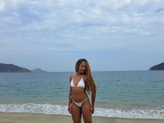 EmmaLatin with long, wavy blonde hair, white bikini, tan skin, small tits, slim, standing on beach with ocean and cloudy sky, arms by sides, looking down.