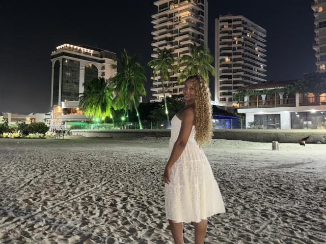 EmmaLatin with long curly blonde hair in a white, textured, sleeveless dress, standing on a sandy beach at night, looking back with her ass slightly raised. Tall buildings.