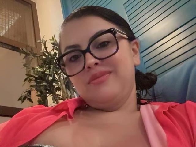 CharlotteDevereaux with glasses, pink shirt, and pink bra, showing cleavage, smiling slightly, dark hair in bun, fair skin, blue background, indoor setting.